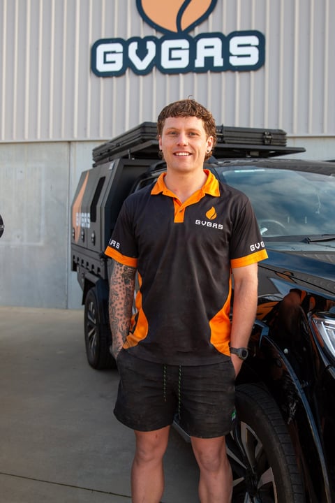 Man wearing black and orange GVGAS polo shirt standing in front of black vehicle and company logo