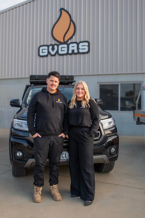 Two people in black GVERAS branded clothing stand in front of a black truck with roof rack at a corrugated metal building with GVERAS logo